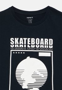 Majica tamnoplave boje od pamuka, s bijelim grafikonom koji prikazuje tekst skateboarda i siluetu unutar kružnog dizajna na prednjoj strani.