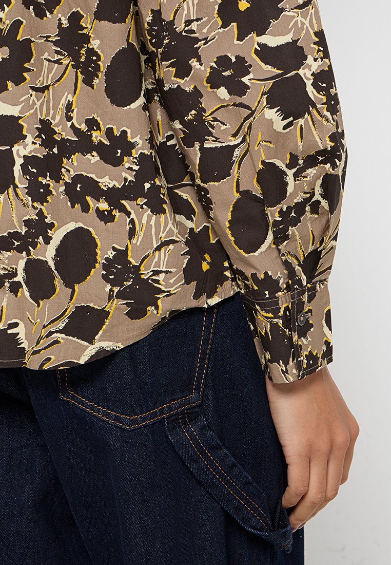 Braun gemustertes Blumenhemd aus leichtem Material mit langen Ärmeln, ausgestattet mit einem Knopfverschluss am Ärmel und einem abgerundeten Saum, kombiniert mit dunklen Jeans.