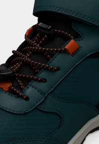 Τεχνητό αθλητικό παπούτσι σε τυρκουάζ χρώμα με υφή στην επιφάνεια, μαύρες λεπτομέρειες, πορτοκαλί εξαρτήματα και μαύρα κορδόνια με πορτοκαλί κλωστές για επιπλέον λεπτομέρεια.