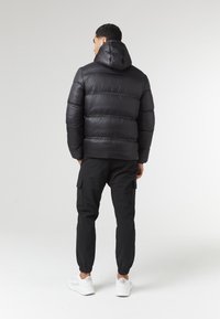 Giacca a vento nera con cappuccio, caratterizzata da sezioni trapuntate e polsini elasticizzati, abbinata a pantaloni cargo neri e sneakers bianche.