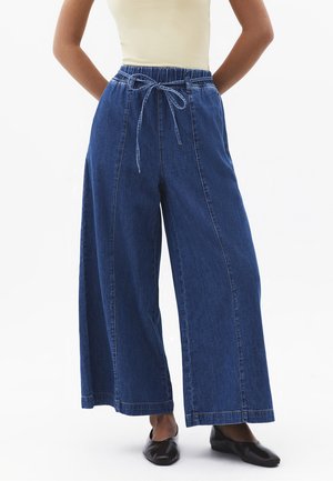 Personne portant un pantalon en denim bleu taille haute à jambes larges avec une ceinture nouée et des chaussures plates noires, debout devant un fond blanc.