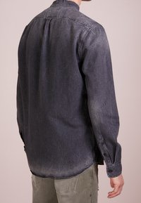 Camisa de denim en gris oscuro con textura desgastada, mangas largas, puños con botones, detalle de pliegue en la espalda y dobladillo curvado.