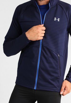 Mann trägt dunkelblaue, mit Reißverschluss versehene Jacke mit gesteppter Brustpartie und Under Armour-Logo, teilweise geschlossen, Hände justieren Reißverschluss und Saum.