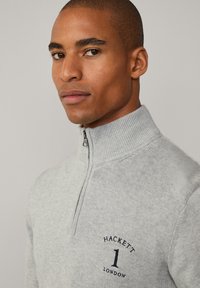 Suéter gris con cremallera en el cuello, hecho de tejido de punto, que presenta un cuello acanalado y un logo bordado en azul marino en el pecho. Textura suave, diseño ajustado.