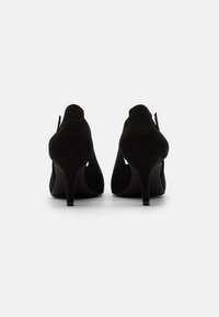Mustat mokkanahka korkokengät, joissa on kaareva muotoilu, kompakti stiletto-korko ja avoin tausta. Sileä pinta, jossa on minimaalinen yksityiskohtina.