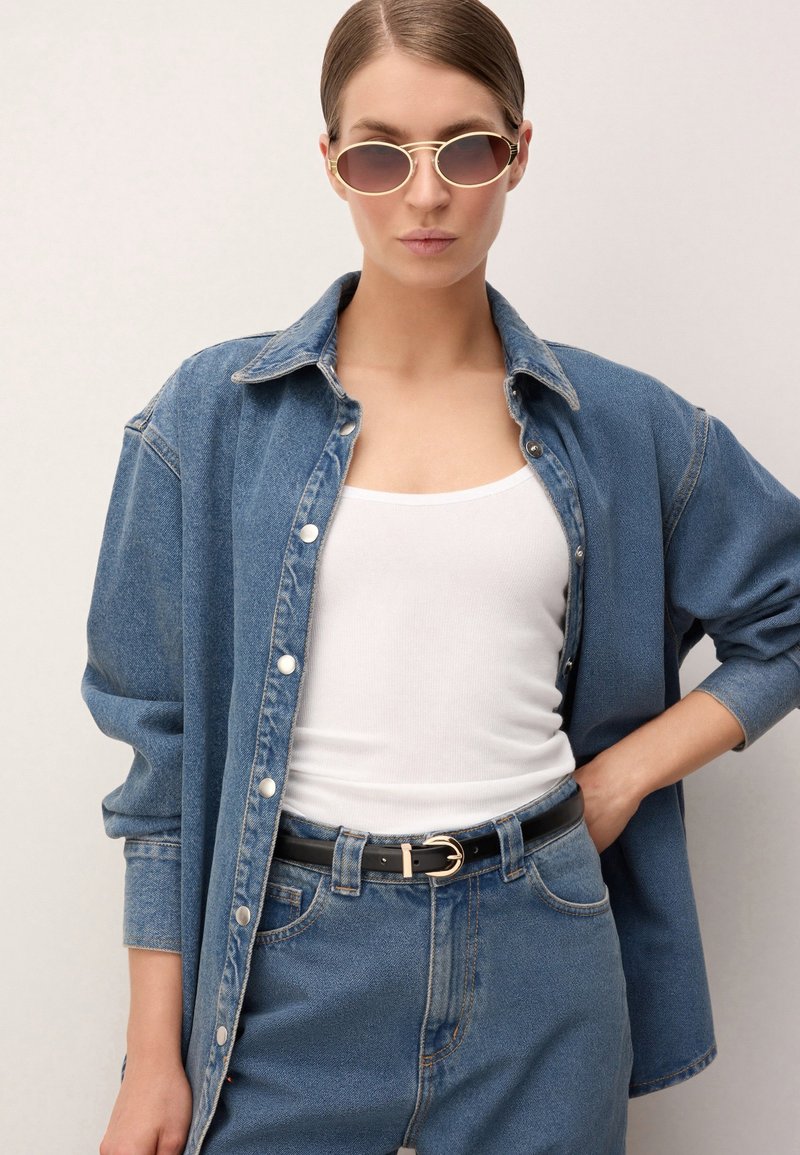 Kobieta w owalnych okularach przeciwsłonecznych, białym topie na ramiączkach, oversize'owej kurtce dżinsowej, jeansach z wysokim stanem i czarnym pasku ze złotą klamrą, z ręką na biodrze.