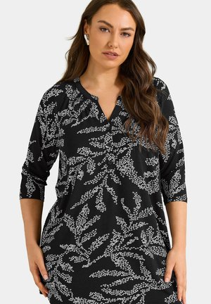 Mujer con vestido negro con patrones florales blancos, mangas tres cuartos y escote en V, de pie frente a un fondo claro y liso.