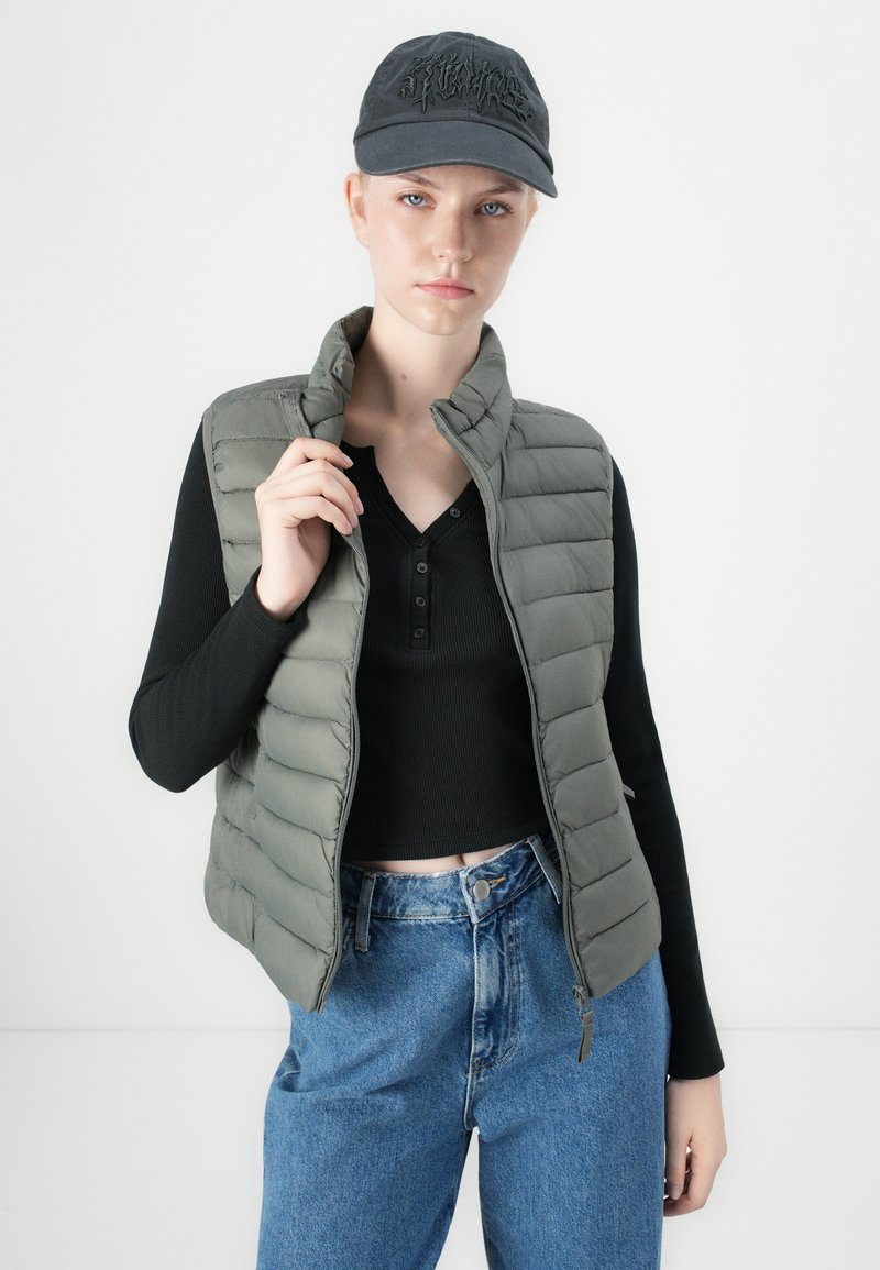 Hellgrüne Steppweste mit hohem Kragen, getragen über einem schwarzen Langarmshirt. Kombiniert mit blauen hoch geschnittenen Jeans und einer dunklen Mütze.