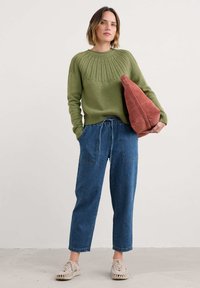 Pull en tricot vert olive avec un empiècement texturé, assorti à un jean en denim bleu clair ample avec poches avant et taille à cordon. Sac en daim couleur rouille.