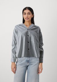 Striped button-up shirt in black and white, featuring a chest pocket, soft fabric, wide sleeves, and a collared neckline. Worn with light jeans.