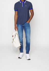 Námořnické polo tričko s červenými a modrými akcenty na límci, doplněné světle modrými zúženými džíny. Drží krémový batoh, na nohou má bílé tenisky.