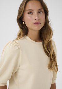 Beige huvudtröja med puffärmar, rund halsringning och slät struktur. Bärs med cirkulära guldörhängen, vilket framhäver den avslappnade stilen.