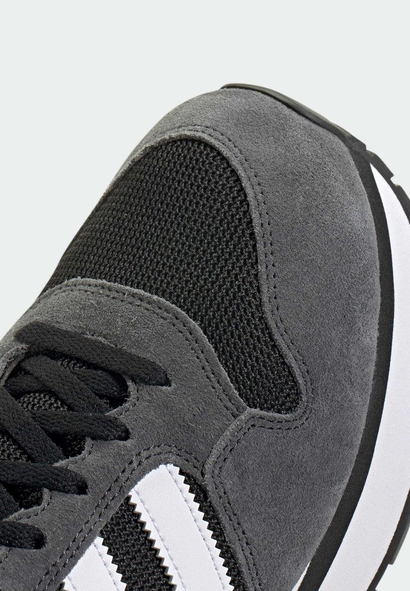 Sneaker grigia che presenta un mix di materiali in camoscio e mesh, lacci neri, strisce bianche e una suola in gomma testurizzata con una forma arrotondata.