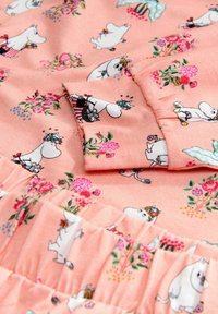 Ferskenfarvet stof med Moomin-illustrationer og blomsterprint, med en flæsekant og blød tekstur. Den detaljerede design forbedrer det visuelle udseende.