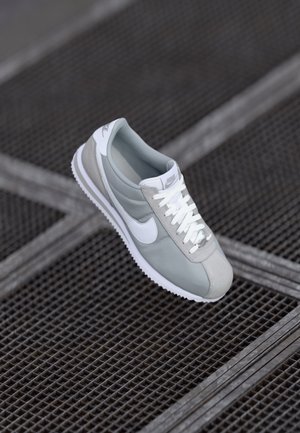 Sive Nike teniske z gladkim zgornjim delom, belimi detajli, teksturiranimi prekrivali in gumijastim podplatom z zigzag vzorcem. vezalke so bele.