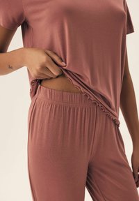 Różowy zestaw odzieży domowej wykonany z miękkiego, gładkiego materiału; bluzka ma falbankowy dół, a spodnie posiadają elastyczny pas.