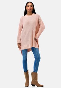 Frau trägt einen oversized pinken Strickpullover, blaue Skinny-Jeans und braune Wildlederstiefel bis zur Mitte der Waden vor einem weißen Hintergrund.