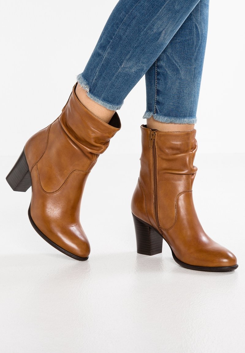 Bottes en cuir marron montant jusqu'au milieu du mollet avec talons carrés, portées avec un jean bleu à ourlet effiloché, pieds croisés et légèrement relevés du sol.