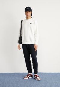 Valkoinen huppari, jossa on musta Nike-logo, musta lippis, mustat collegehousut ja punavalkoiset Nike-tennarit, henkilön päällä, joka seisoo vaaleaa taustaa vasten.