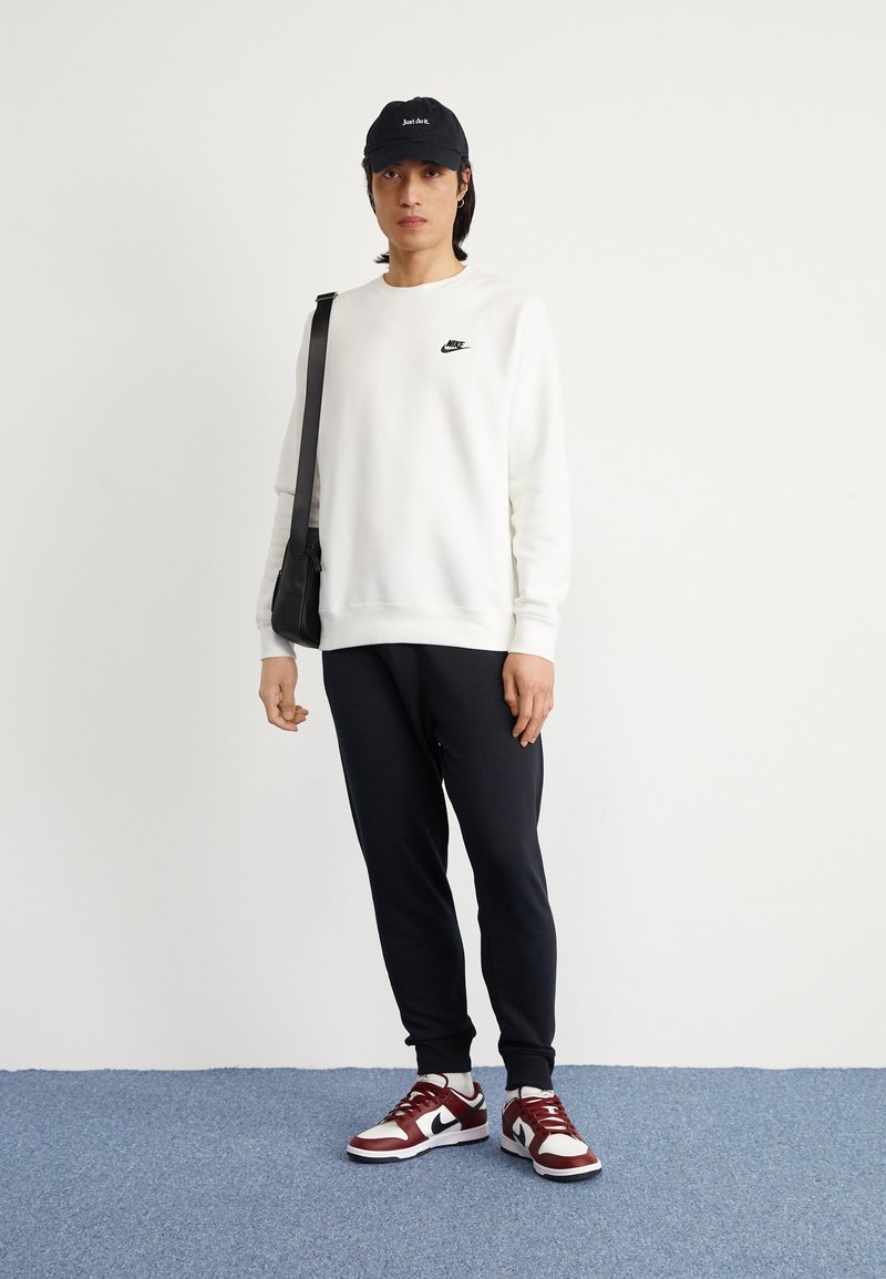 Sudadera blanca con el logo negro de Nike, gorra negra, pantalones de chándal negros y zapatillas Nike rojas y blancas, en una persona de pie frente a un fondo claro.