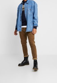 Denim jacket with a relaxed fit, blue color, two front pockets. Layered over a navy sweater with a geometric pattern. Brown corduroy pants and black lace-up boots.