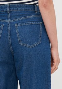 Ciemnoniebieskie dżinsy o wysokim stanie, z dwoma tylnymi kieszeniami i widocznym przeszyciem. Tkanina wydaje się lekko teksturowana.