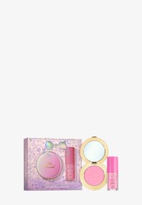 Te mooie roze blush compact van Too Faced met spiegel, naast een roze lipgloss tube, allemaal in een versierde paarse doos met bloemenpatronen en een strikontwerp.