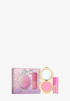 Te mooie roze blush compact van Too Faced met spiegel, naast een roze lipgloss tube, allemaal in een versierde paarse doos met bloemenpatronen en een strikontwerp.