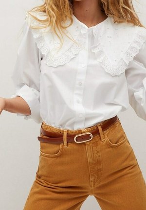 Biała koszula zapinana na guziki z dużym koronkowym kołnierzem, włożona do wysokich musztardowych spodni z brązowym paskiem.