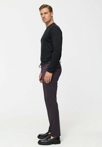 Man stands sideways wearing black long-sleeve shirt, dark purple drawstring pants, and black loafers against a plain white background.