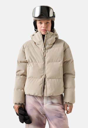 Person trägt eine beige gesteppte Jacke mit hohem Kragen, hellrosa Skihose, Skihelm, Skibrille und hält schwarze Handschuhe in einer Hand.