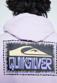 Lila hoodie med en stor svart och gul grafik med "Quiksilver"-logotypen, omgiven av lila med taggiga kanter.
