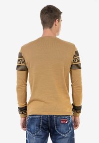Beige Pullover mit langen Ärmeln, mit schwarzen gemusterten Akzenten an den Schultern. Getragen mit blauen Jeans, die eine strukturierte Oberfläche zeigen.