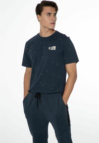 Marineblaues T-Shirt mit kurzen Ärmeln, das mit bunten Punkten verziert ist und das "NXG"-Logo auf der Vorderseite trägt. Hergestellt aus einem weichen Stoff mit lockerer Passform.