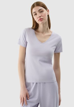 Jonge vrouw met stijl rood haar, die een lichtpaars T-shirt met korte mouwen en een V-hals draagt, en een bijpassende broek tegen een effen achtergrond.