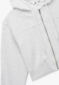Felpa con cappuccio grigia chiara, rifinita e realizzata in un tessuto morbido. Presenta una zip frontale, lacci e polsini a coste con cuciture oblique.
