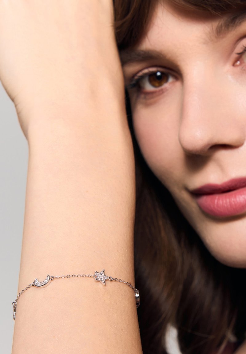 Srebrna bransoletka łańcuszkowa z motywem półksiężyca i gwiazdy, ozdobiona małymi przezroczystymi kamieniami szlachetnymi, noszona na jasnej karnacji.