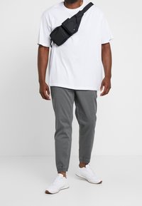 Valkoinen lyhythihainen t-paita, harmaat kapenevat housut ja valkoiset tennarit. Musta olkalaukku, jossa on kaksi osastoa, kannetaan olkapään yli.