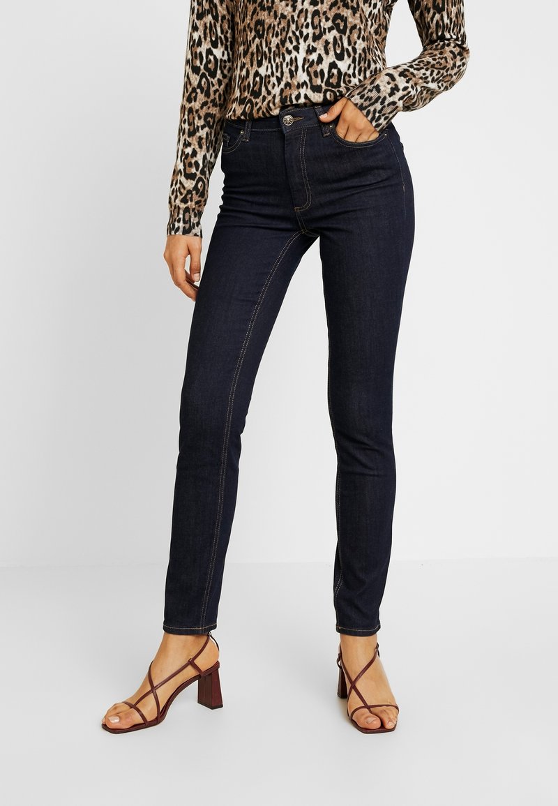Femme portant un jean skinny foncé, un haut à manches longues imprimé léopard, et des sandales à talons marron à lanières, debout avec une main dans la poche.