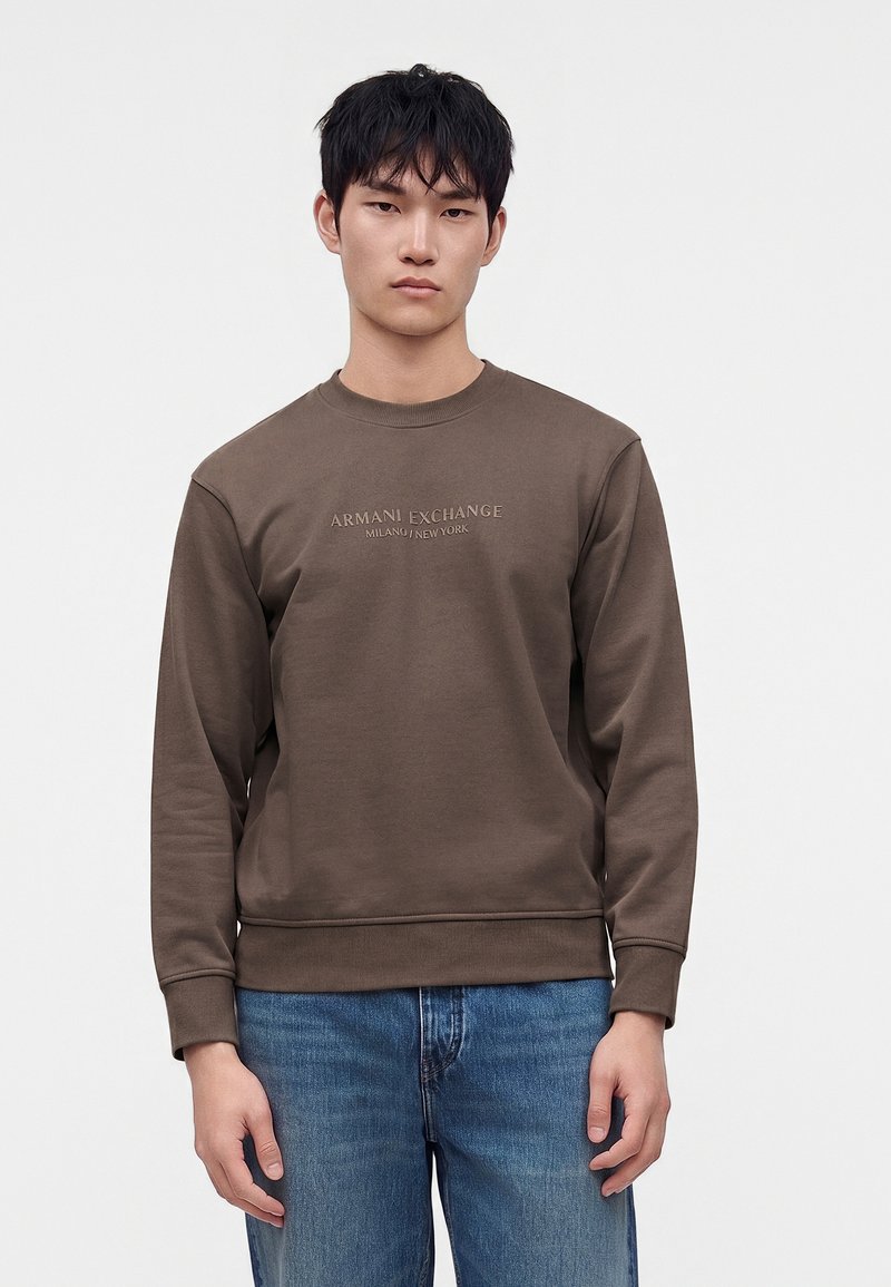 Νέος άνδρας που φοράει ένα καφέ φούτερ Armani Exchange και μπλε τζιν, στέκεται μπροστά σε ένα απλό φωτεινό φόντο.