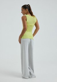 Mujer de pie de lado, vestida con una camiseta sin mangas de color verde claro y pantalones grises sueltos, con el pelo recogido en una cola de caballo.