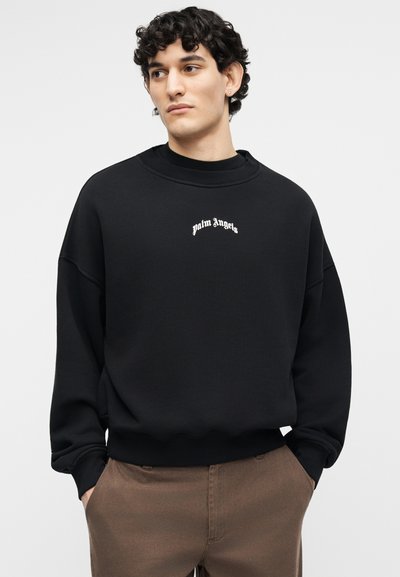 Νεαρός άνδρας με σγουρά μαλλιά, φοράει μαύρο φούτερ με το κείμενο "palm angels" και καφέ παντελόνι, με τα χέρια στις τσέπες, κοιτάζοντας πλάγια.