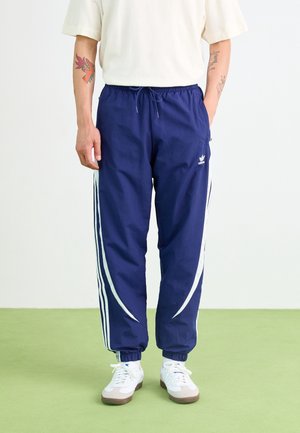 Personne portant un pantalon de survêtement Adidas bleu marine avec des bandes blanches et des baskets blanches, debout sur un sol vert clair devant un fond blanc.