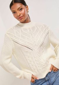 Gestrickter cremefarbener Pullover mit hohem Kragen und aufwendigen Zopfmustern, strukturierten Ärmeln und gerippten Bündchen, kombiniert mit blauen Jeans.