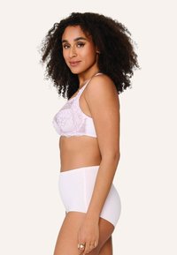 Kanten en gaas bralette in zachte roze met verstelbare bandjes; gecombineerd met hoge taille witte onderbroek. Model in zijprofiel tegen lichte achtergrond.