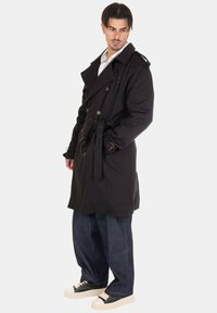 Cappotto nero a doppio petto con bottoni, vita fermata da una cintura e spalline, abbinato a jeans blu larghi e scarpe da ginnastica nere.