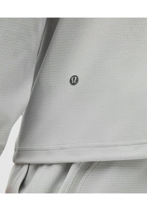 Ljusgrå atletisk topp med strukturerad tyg, rund halsringning och en liten logotyp på vänster sida. Synliga sömmar och lagerdesign.