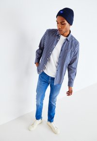 Blau-weiße kariierte Hemd, weißes T-Shirt darunter, blaue Jeans, weiße Sneaker und eine marineblaue Mütze mit einem Logo. Einfaches, lässiges Ensemble.