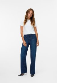 Camiseta blanca de manga corta combinada con unos pantalones vaqueros de pierna ancha azul oscuro, que presentan bolsillos frontales y una textura de denim suave. Tacones de lunares en blanco y negro.
