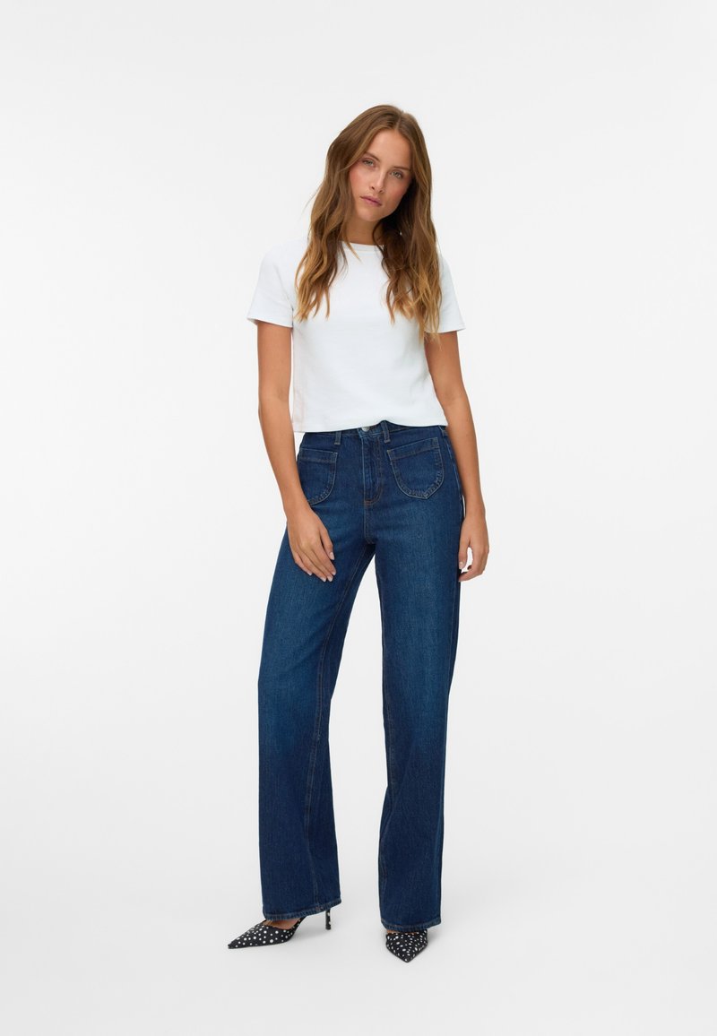 Camiseta blanca de manga corta combinada con unos pantalones vaqueros de pierna ancha azul oscuro, que presentan bolsillos frontales y una textura de denim suave. Tacones de lunares en blanco y negro.